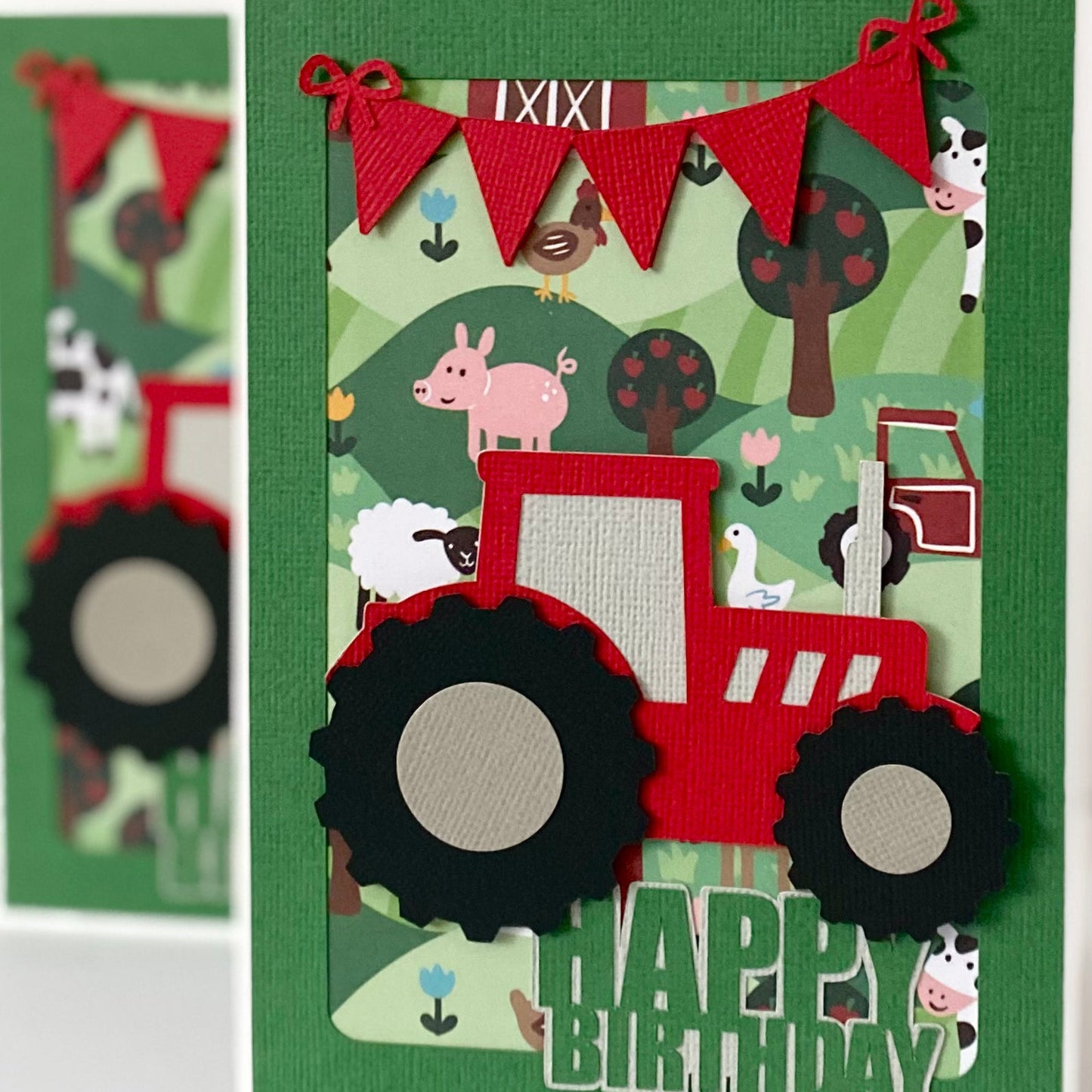 A handmade layered paper card featuring a red tractor with black wheels, a green background with farm animals, and a red bunting banner, with 'HAPPY BIRTHDAY' text at the bottom.